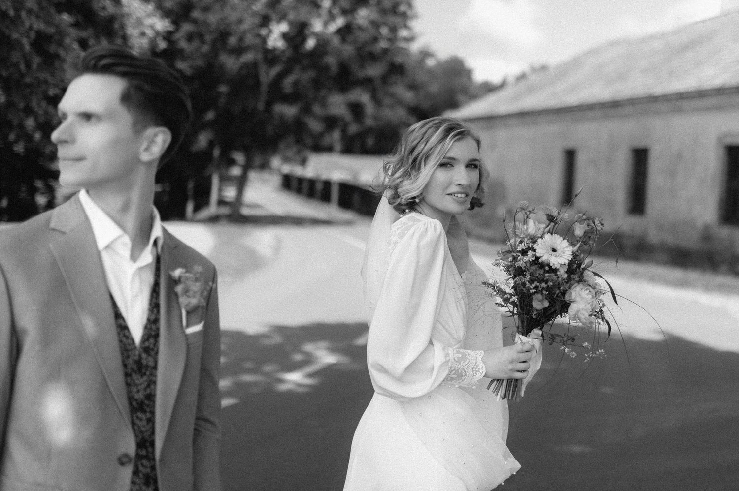 Black and white wedding photo of bride holding bouquet and groom in suit outdoors at elegant venue.