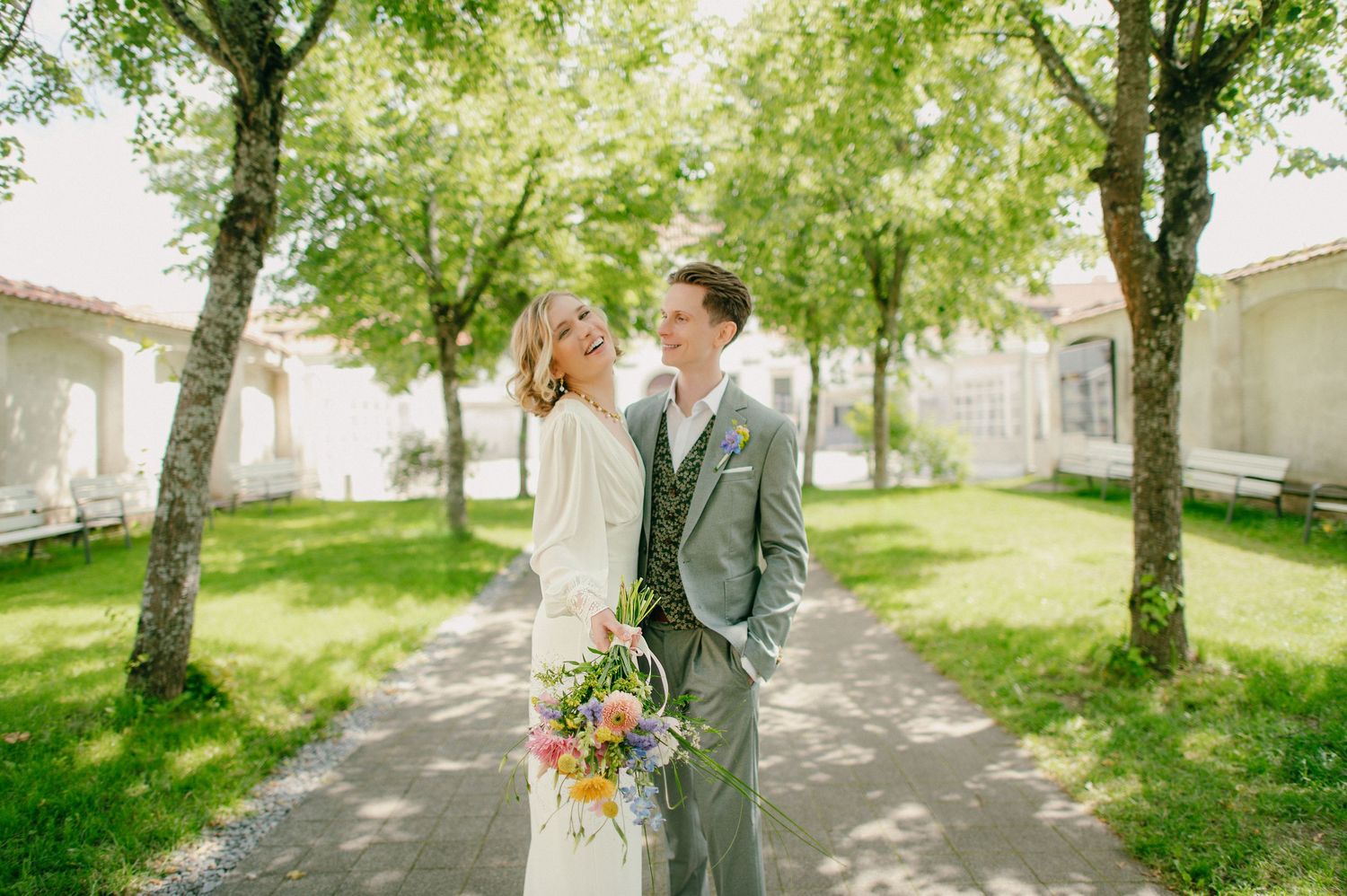 Bride and groom share a romantic moment outdoors, surrounded by lush green trees, as she holds a colorful wedding bouquet.