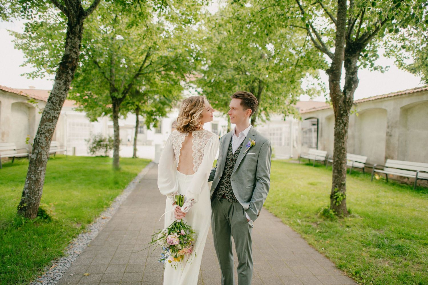 Bride and groom share a romantic moment on a tree-lined path, she in a white gown, he in a gray suit.