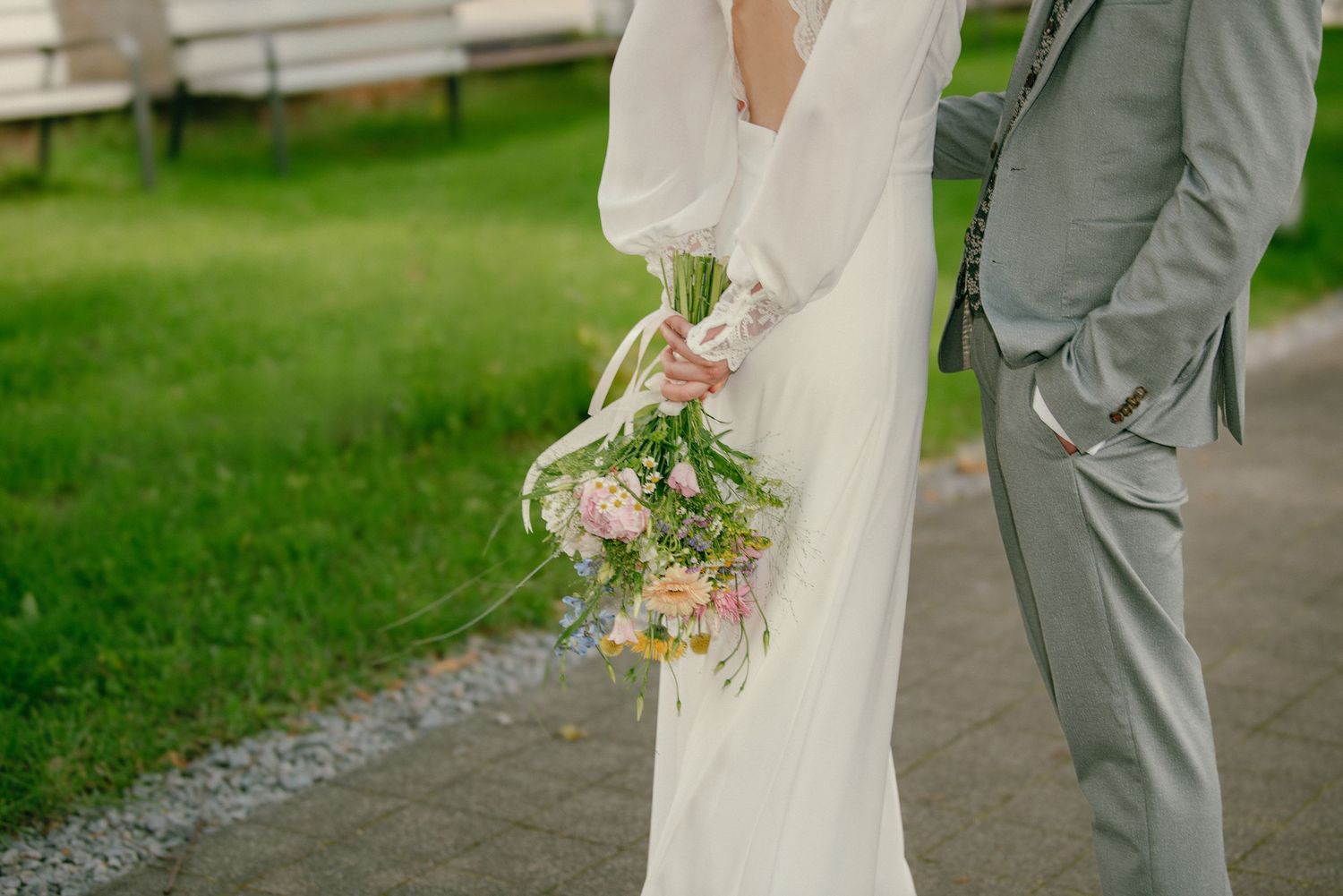 Bride in white gown holding pink wildflower bouquet standing beside groom in gray suit on garden path.