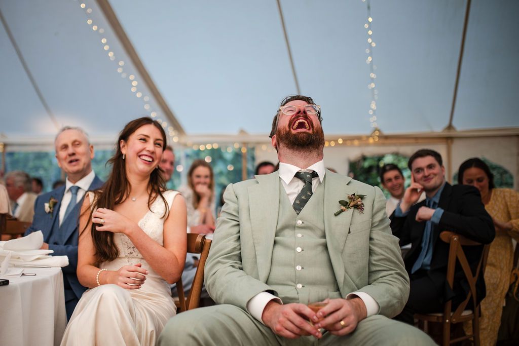 Bride and groom laughing joyfully during wedding reception speeches inside elegant marquee tent.