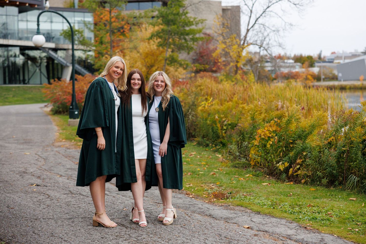 Three women in green graduation gowns smiling outdoors on a campus path surrounded by autumn foliage.
