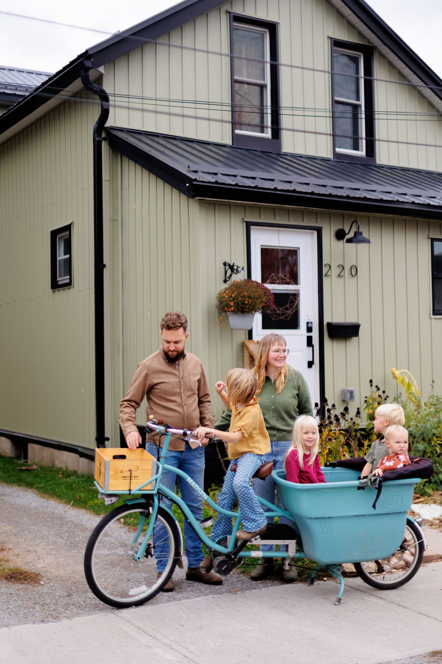 Family with children loading a cargo bike outside a green house numbered 220 on a sunny day.