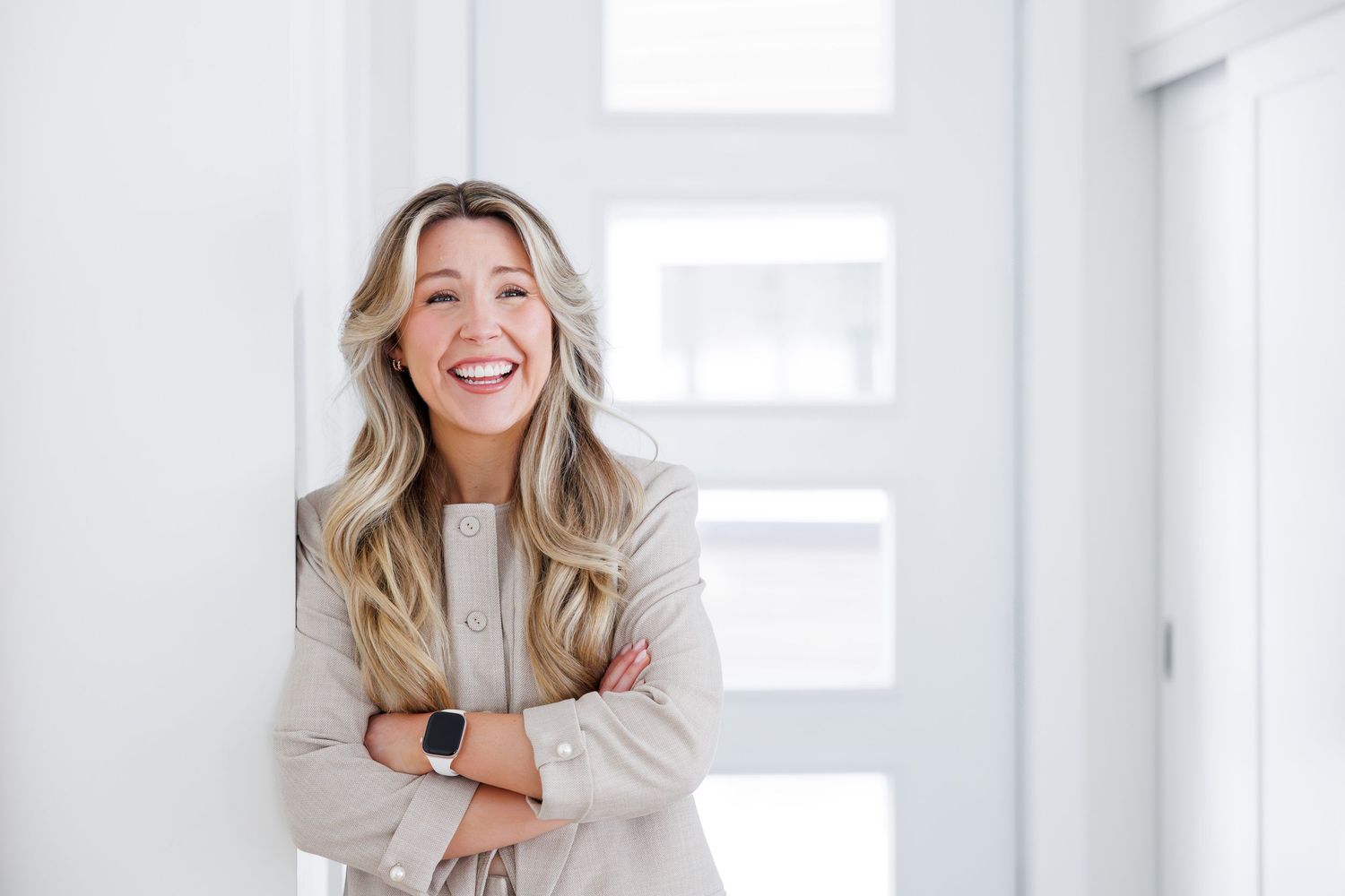 Smiling blonde woman with arms crossed wearing a beige blazer and smartwatch, standing in a bright modern hallway.