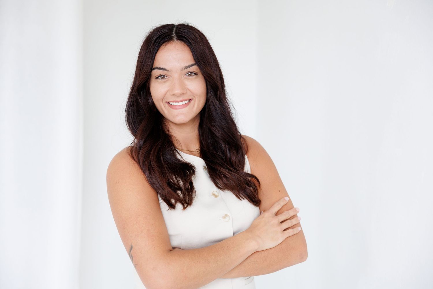 Smiling young woman with dark hair, arms crossed, wearing a white sleeveless top against a bright white background.