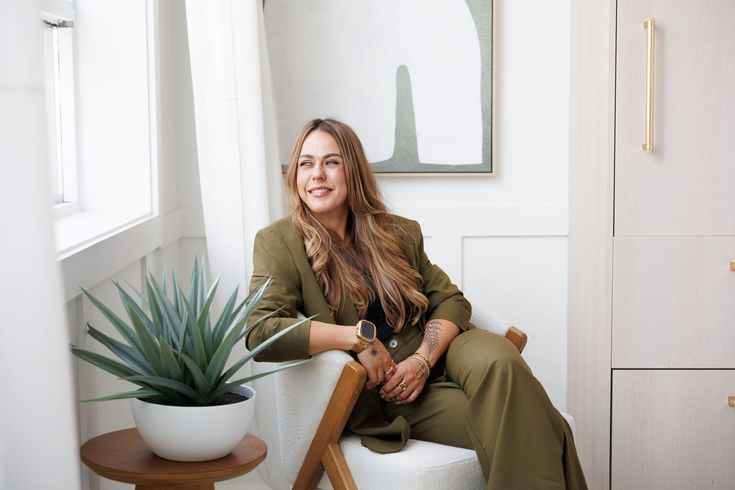 Woman in olive green suit sitting relaxed in bright white room with aloe plant and modern art.