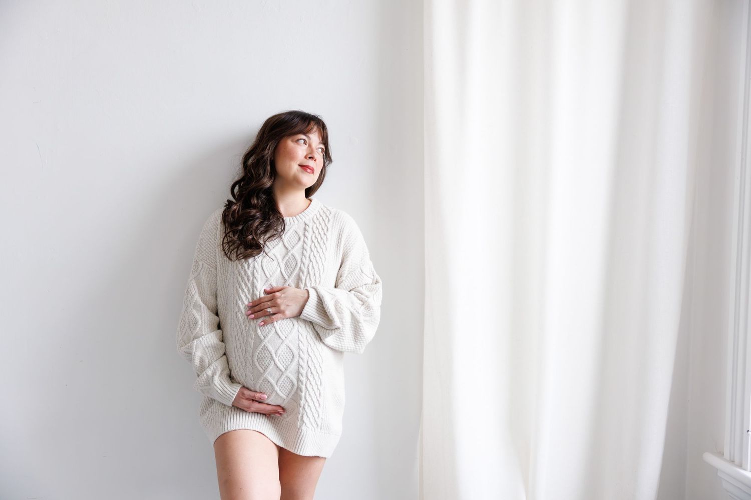 Pregnant woman in cream cable-knit sweater dress cradling baby bump, leaning against white wall near sheer curtains.