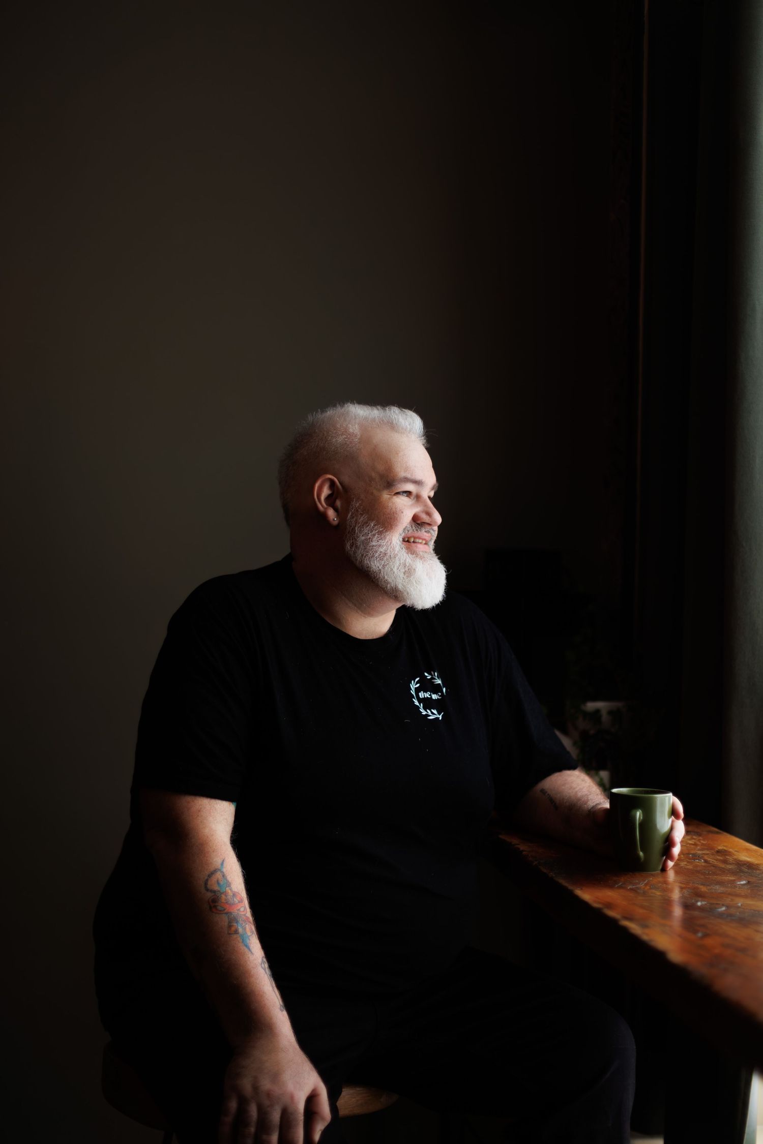 Middle-aged man with white beard and tattoos sits by window holding green mug, wearing black t-shirt in moody lighting.
