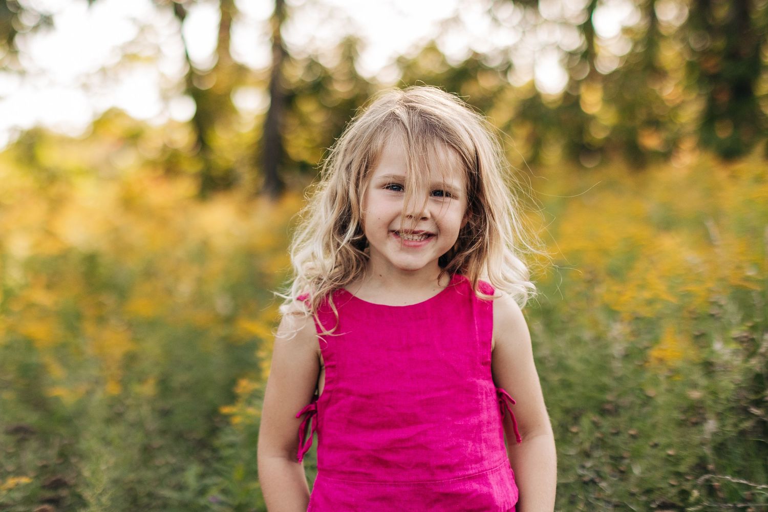 Smiling young blonde girl in pink sleeveless top standing in a sunny outdoor field.
