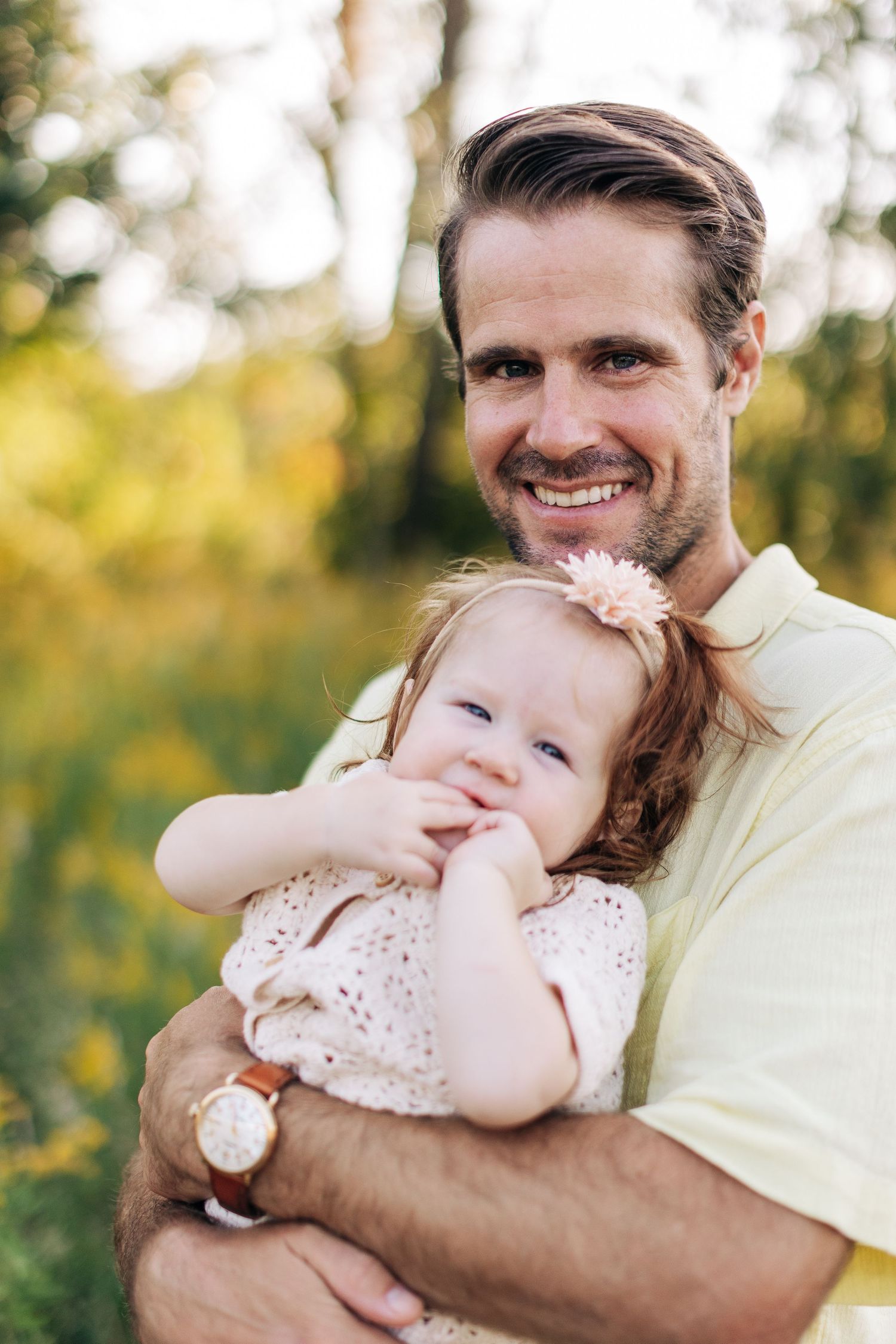 Smiling father in yellow shirt holding cute baby girl with flower headband outdoors.