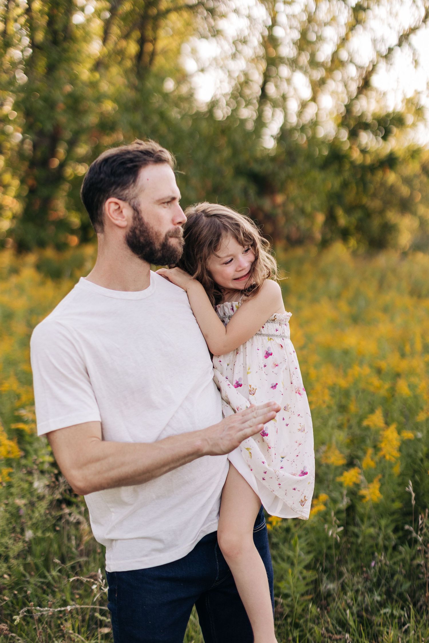A bearded father tenderly holds his young daughter in a golden sunlit field of wildflowers.