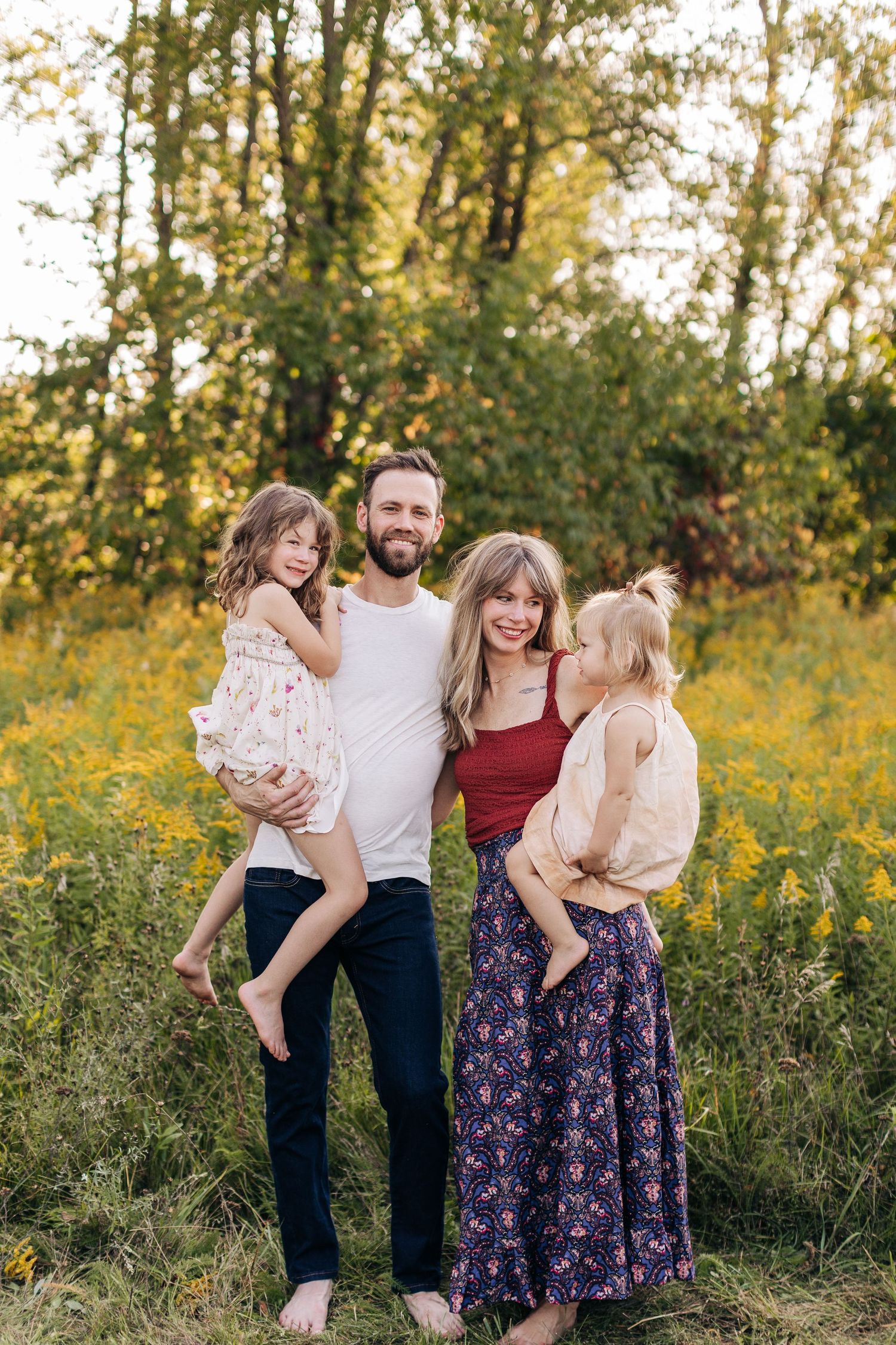 Smiling family of four holding children in a vibrant wildflower field at sunset.