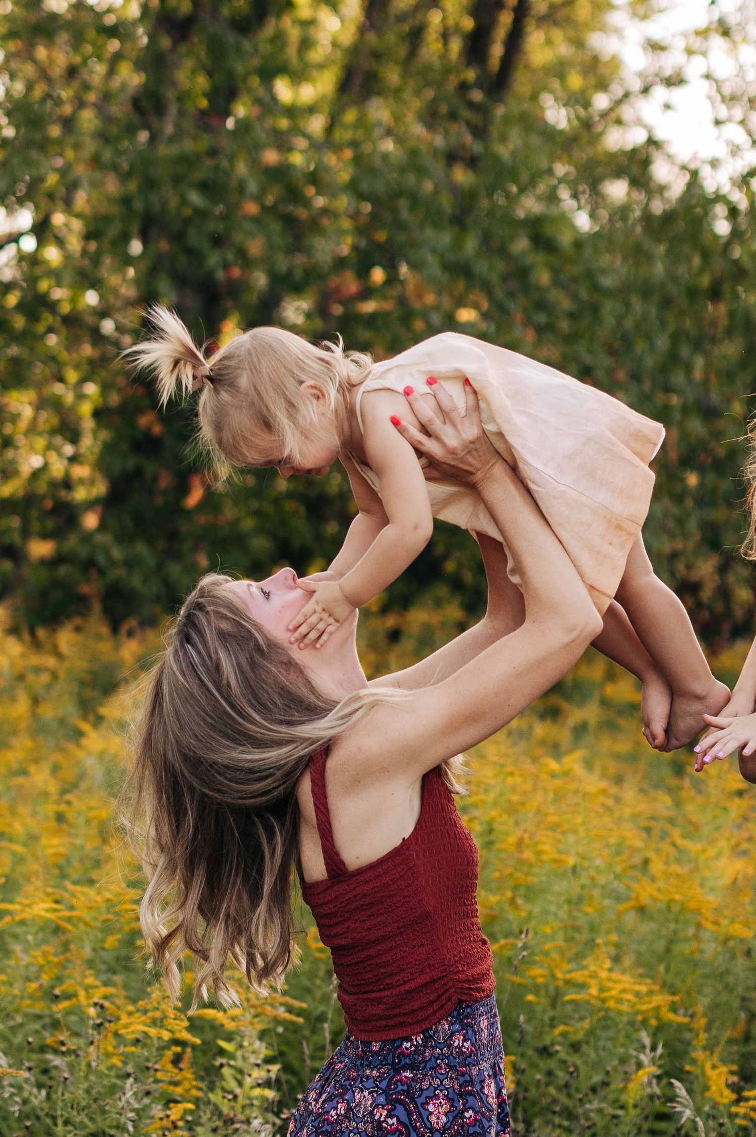 Mother joyfully lifting her smiling toddler daughter in a pink dress above a field of yellow flowers.