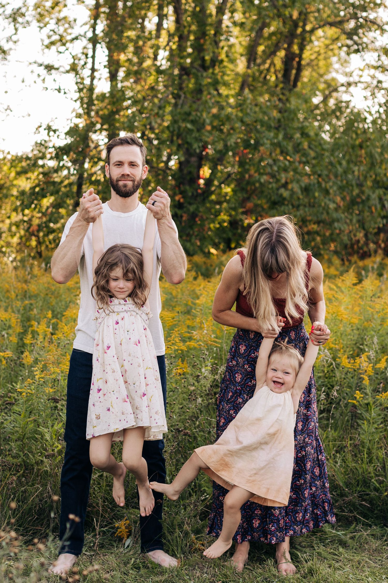 A bearded father and blonde mother playfully lift their two young daughters in a sunny meadow.