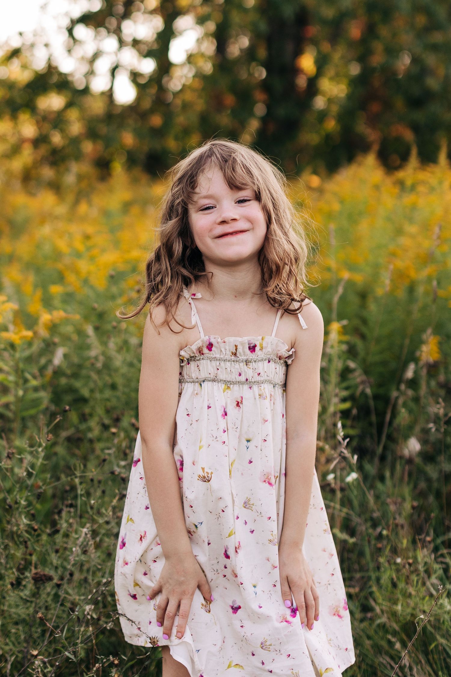 A young girl in a white floral sundress smiles sweetly in front of yellow wildflowers outdoors.