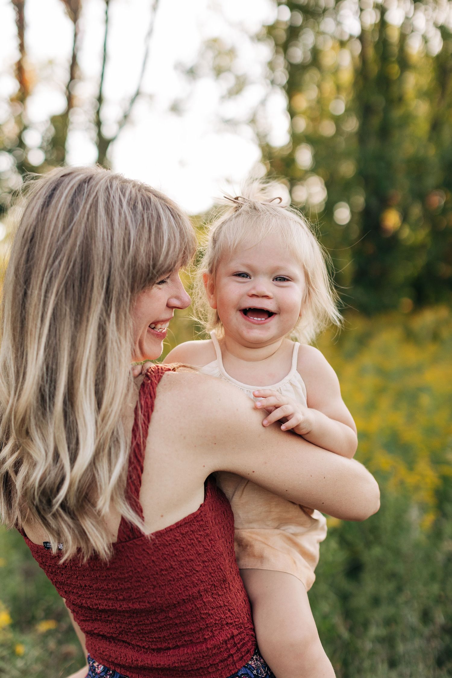 A laughing blonde toddler is held by a smiling woman in a red top in a sunny garden.