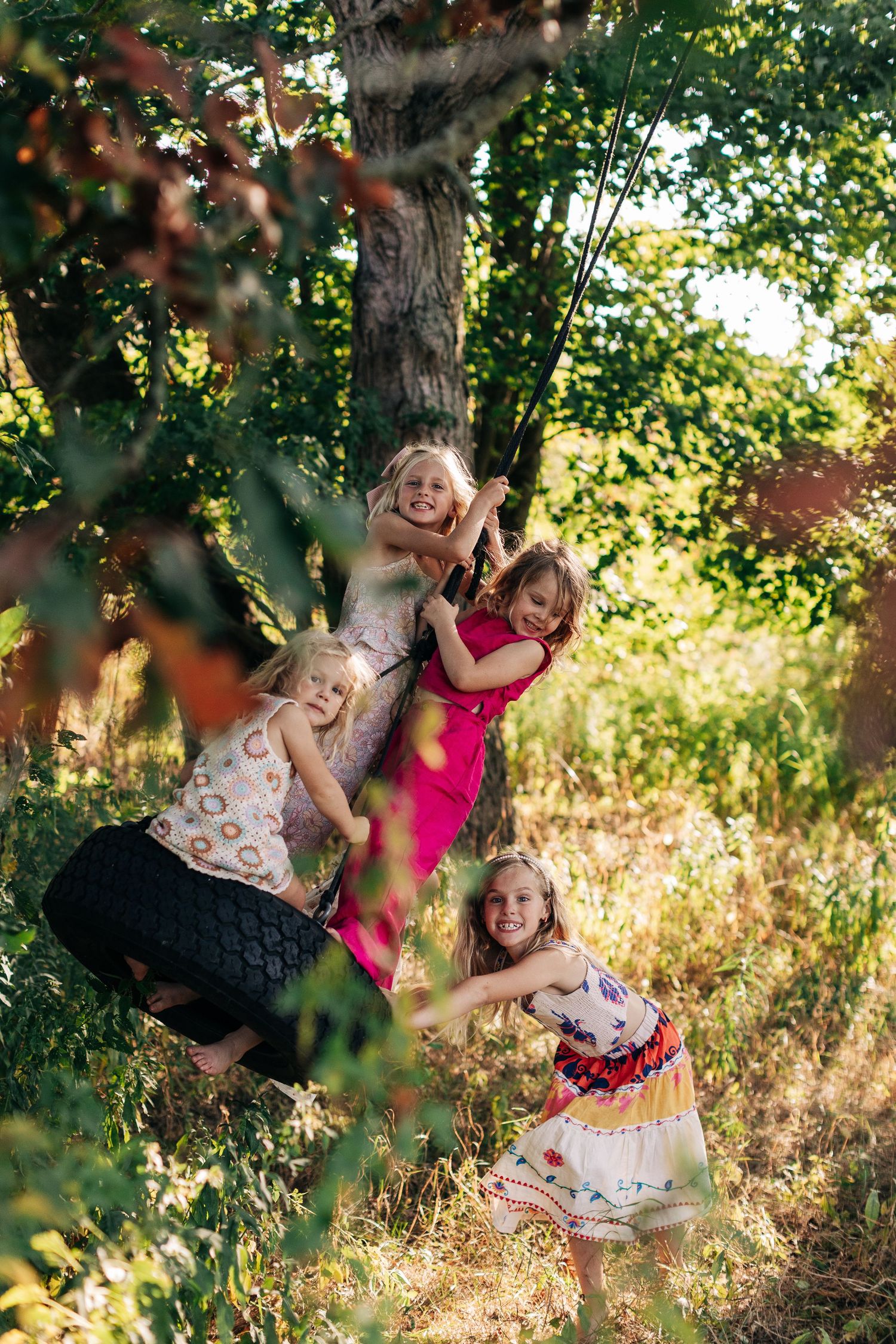 Four young girls playing and laughing on a rope swing tied to a tree outdoors.