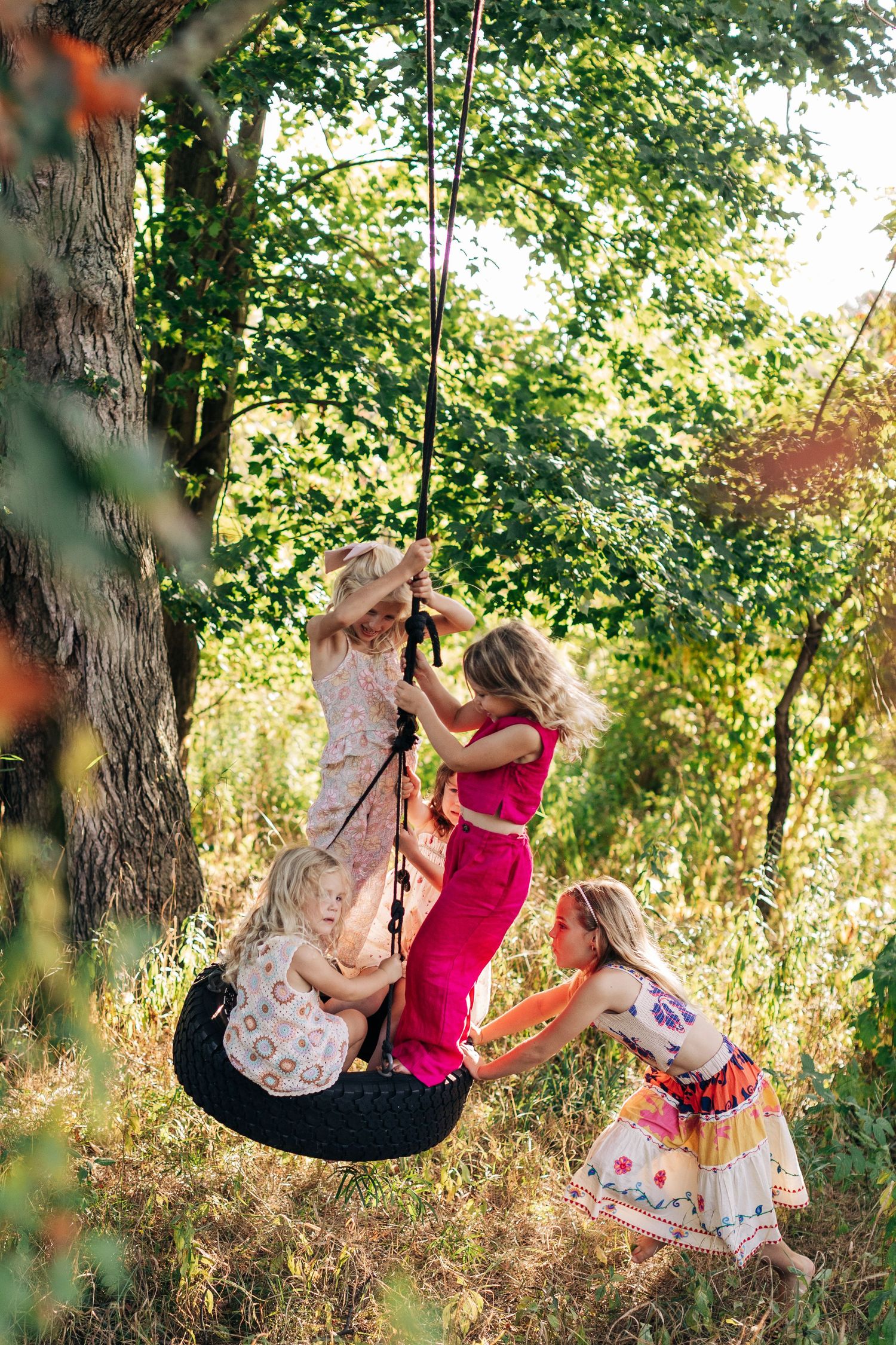 Several young girls in colorful dresses play together on a rope tire swing under a leafy tree.