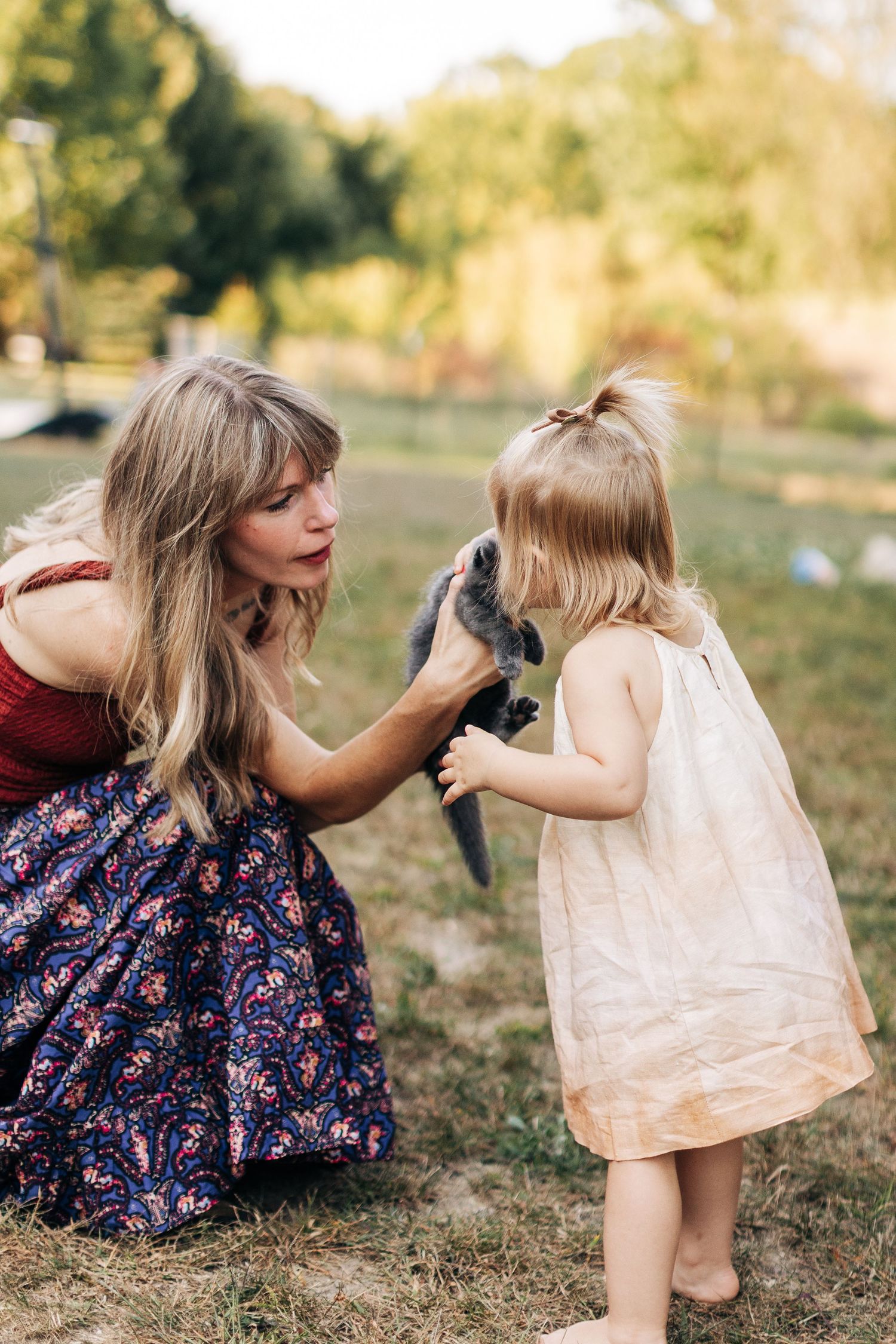 A mother and toddler girl in dresses interact with a small rabbit outdoors in a sunny green park setting.