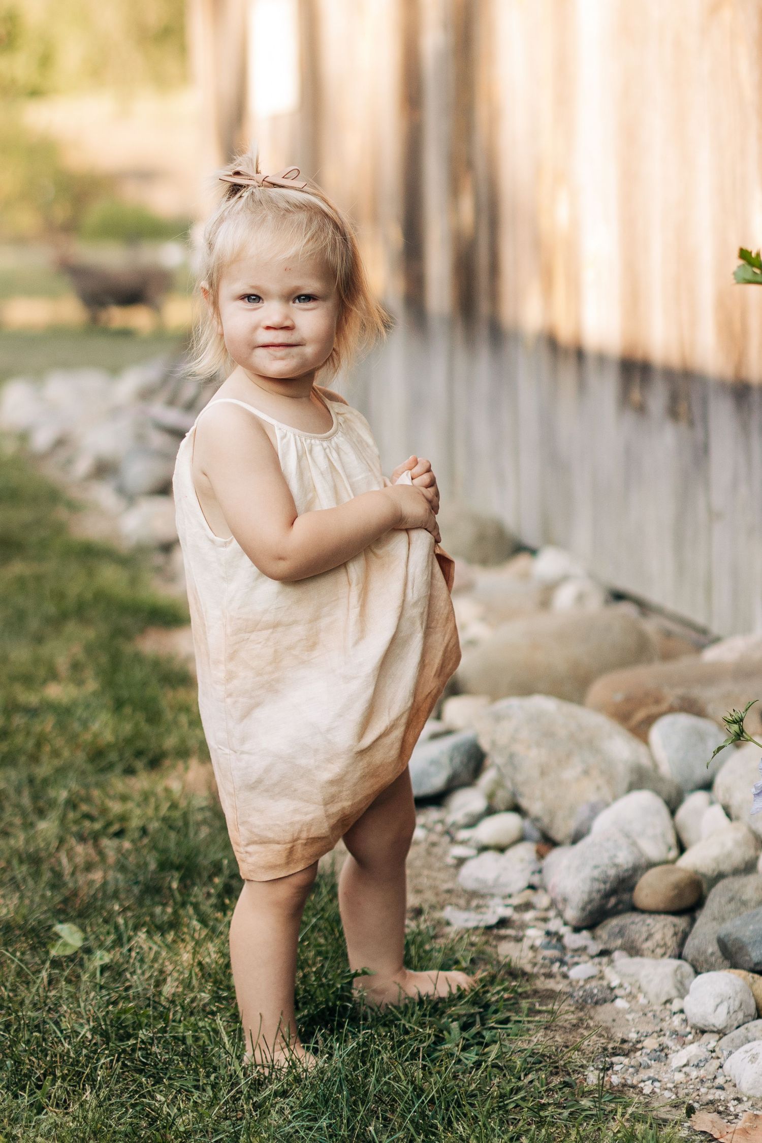 A barefoot toddler girl in a beige linen dress stands on grass beside a wooden fence and stones.