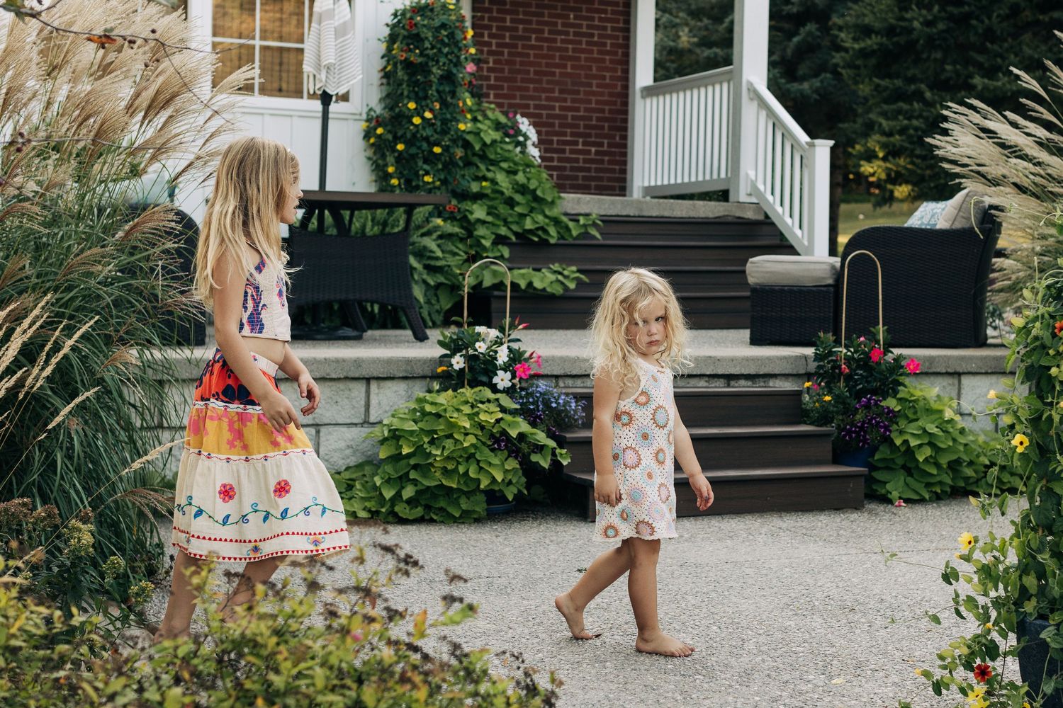Two young girls in floral dresses walking along a garden path in front of a charming brick home.