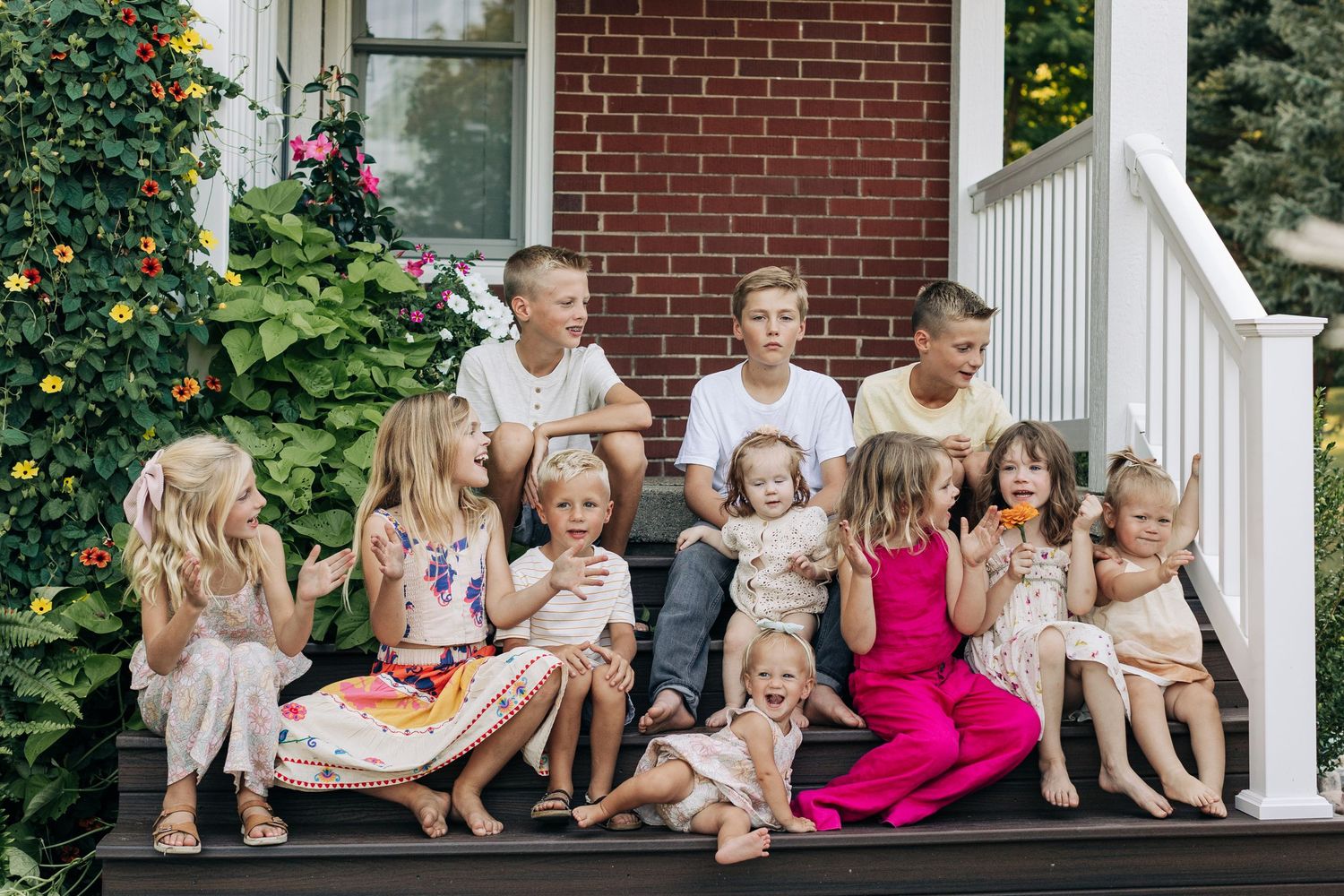 A large group of young children sit and laugh together on a brick home's front porch steps.
