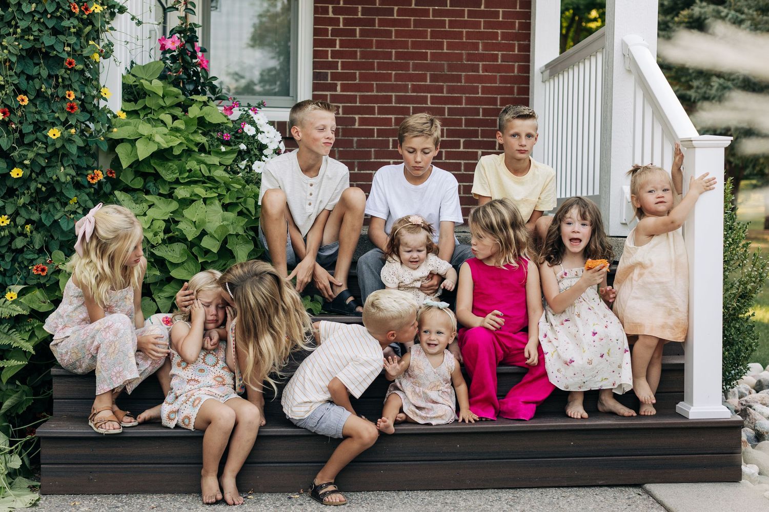 Large group of children sitting together on porch steps surrounded by lush garden flowers.