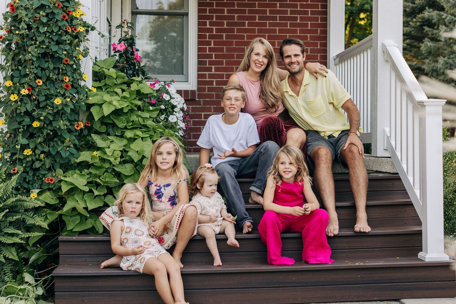 Happy family of seven posing on front porch steps of brick home with colorful flowers.