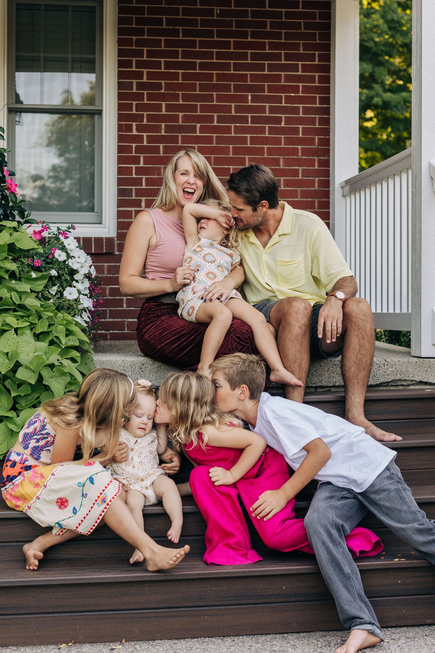 A joyful family with five children laughs together on the front porch steps of a brick home.