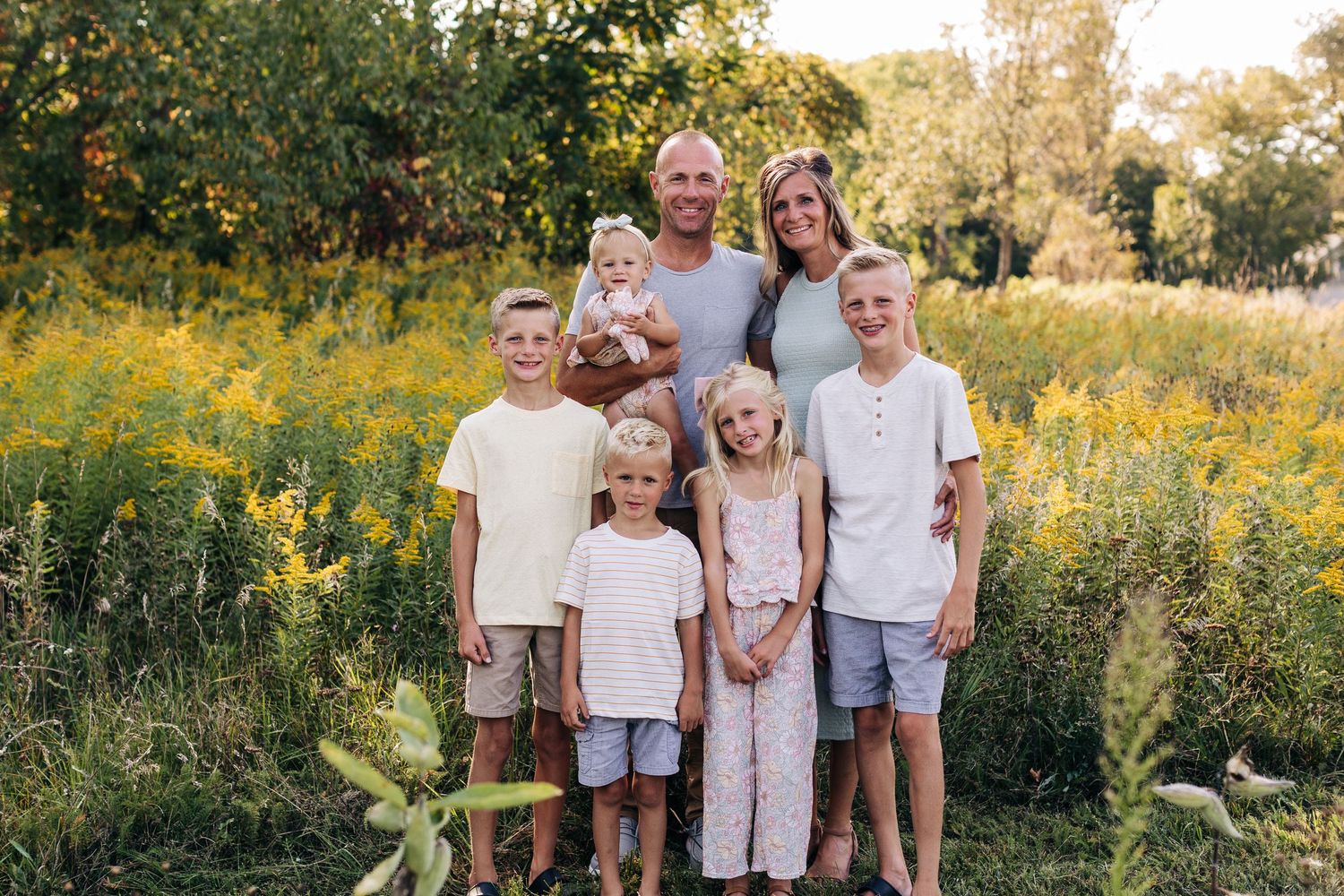 Family of seven posing outdoors in a sunny meadow with yellow wildflowers and green trees in the background.