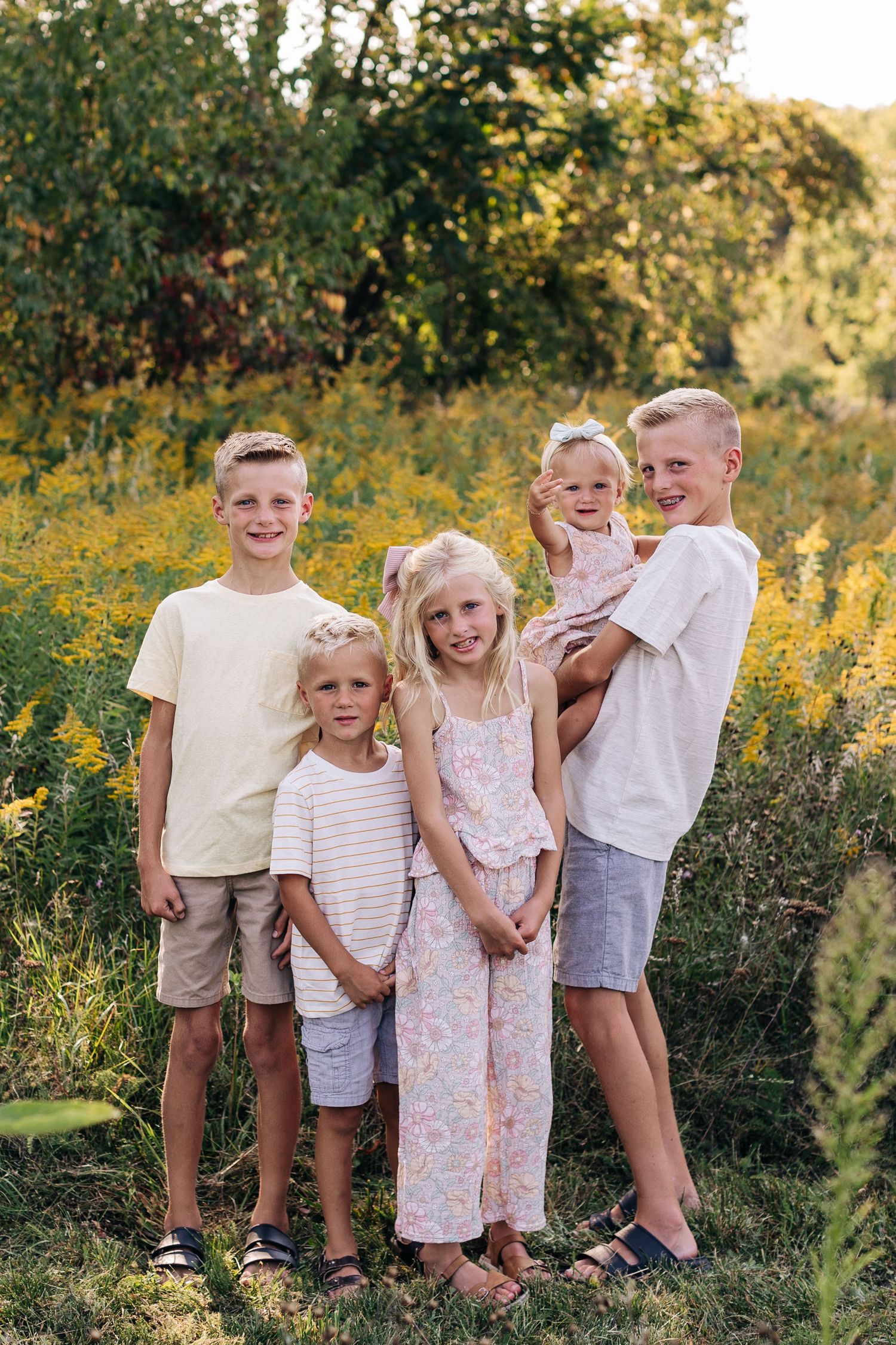 Five blonde siblings pose together in a sunny field of yellow wildflowers during a warm summer day.
