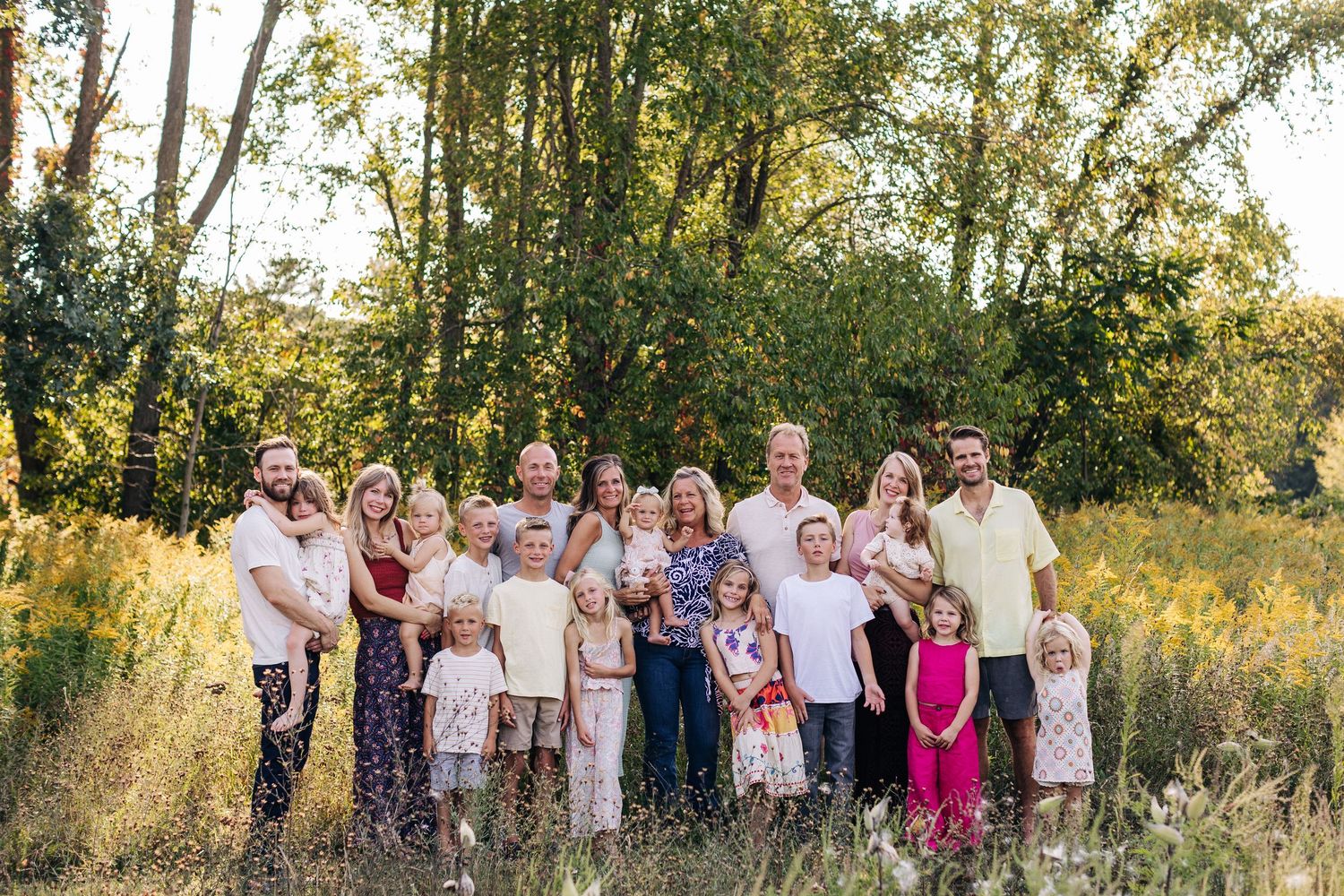 Large extended family posing together outdoors in a sunny meadow surrounded by tall trees and golden wildflowers.