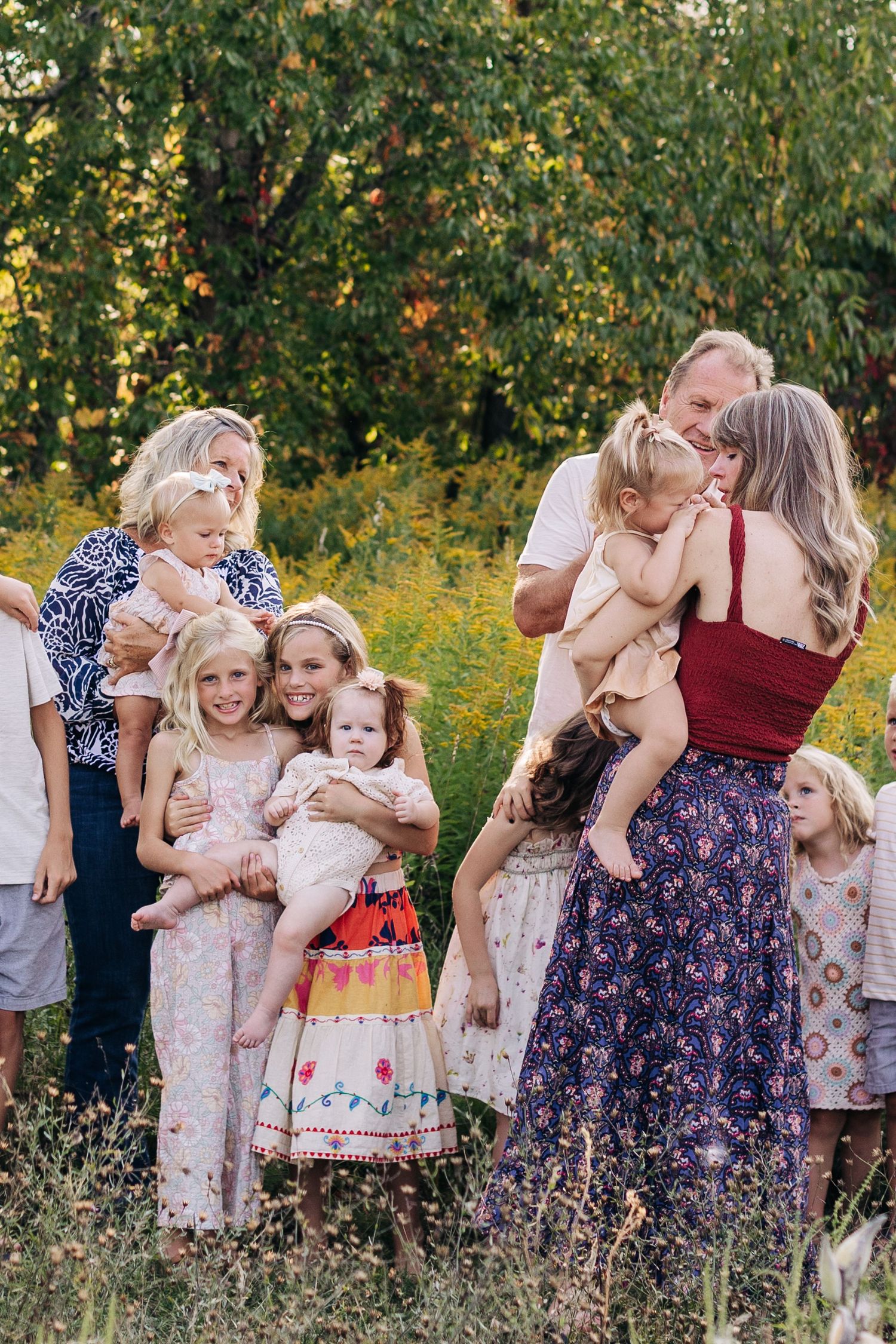 Large family gathering outdoors in a sunny meadow with children being held and hugged among wildflowers.