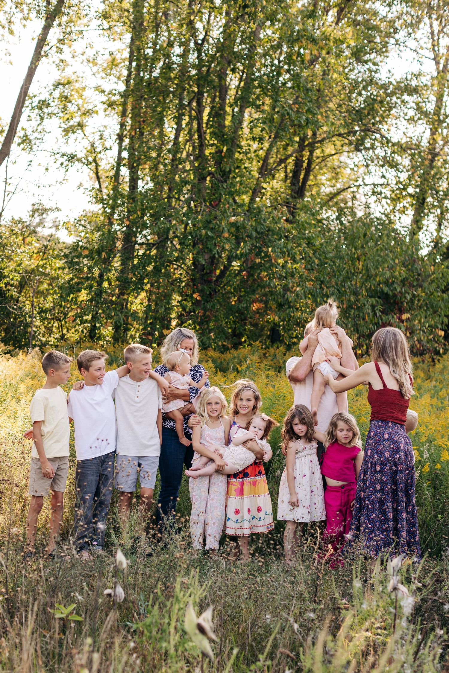 Group of children posing together in a sunlit field with green trees, some holding younger siblings.