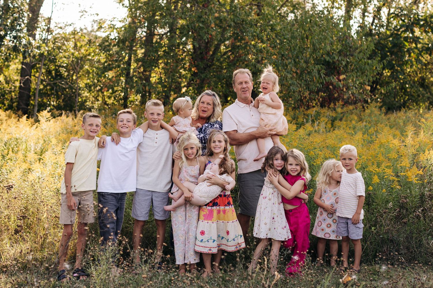 Large family portrait with parents and children posing outdoors in a golden field surrounded by trees.