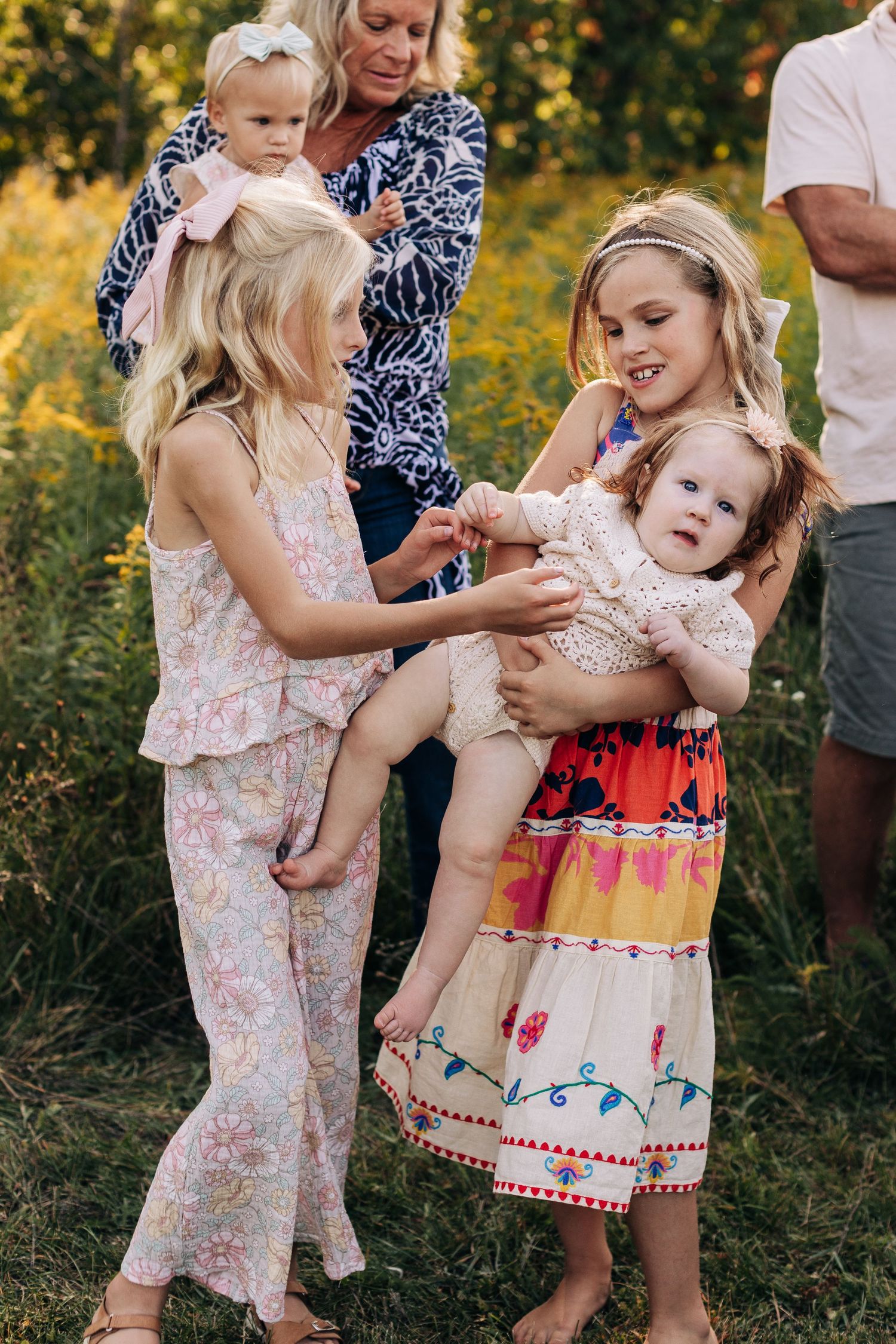 Two girls in floral dresses hold a smiling baby outdoors while a woman in blue stands behind them.