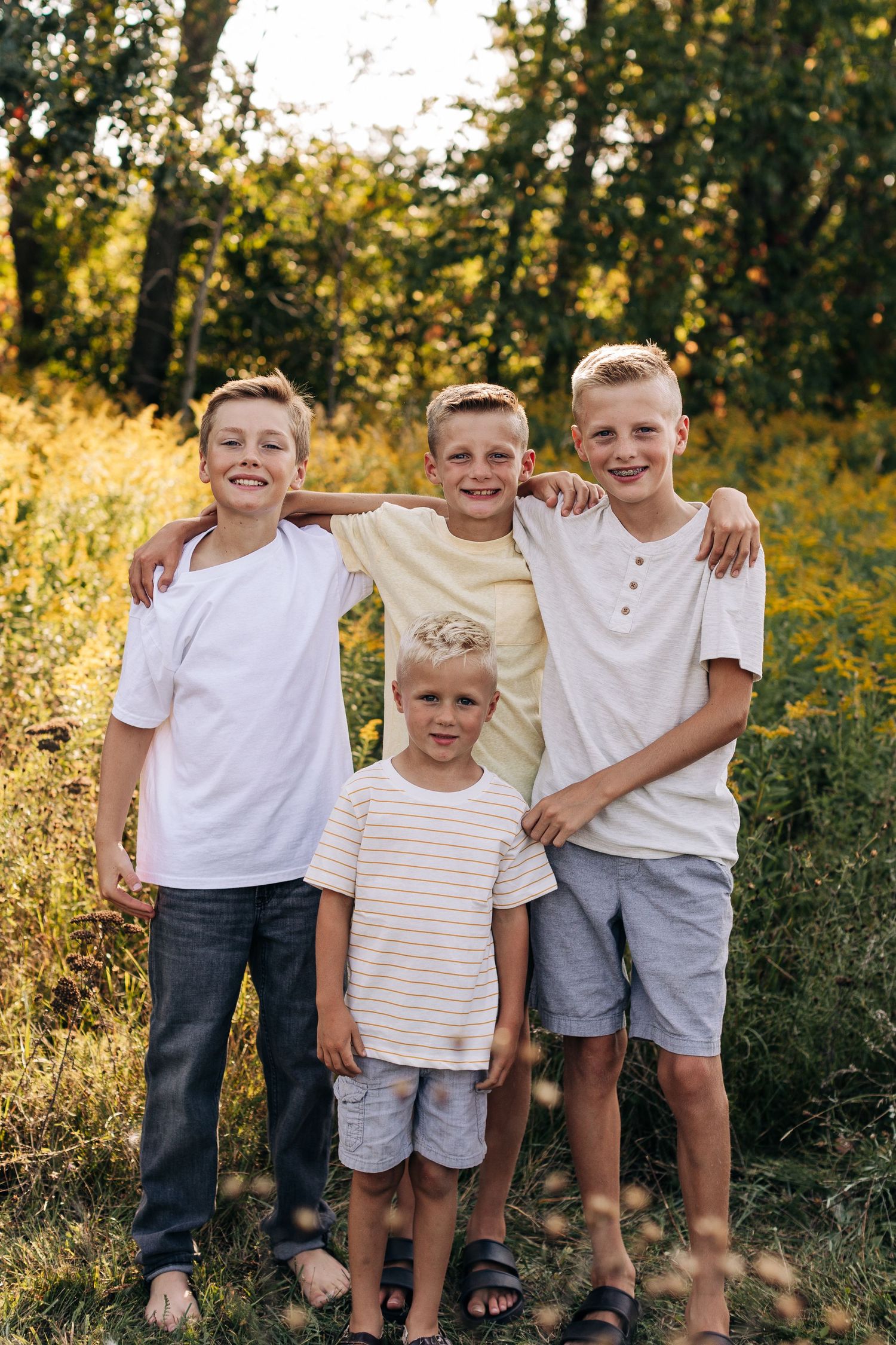 Four smiling blonde brothers pose together with arms around each other in a sunlit outdoor field.
