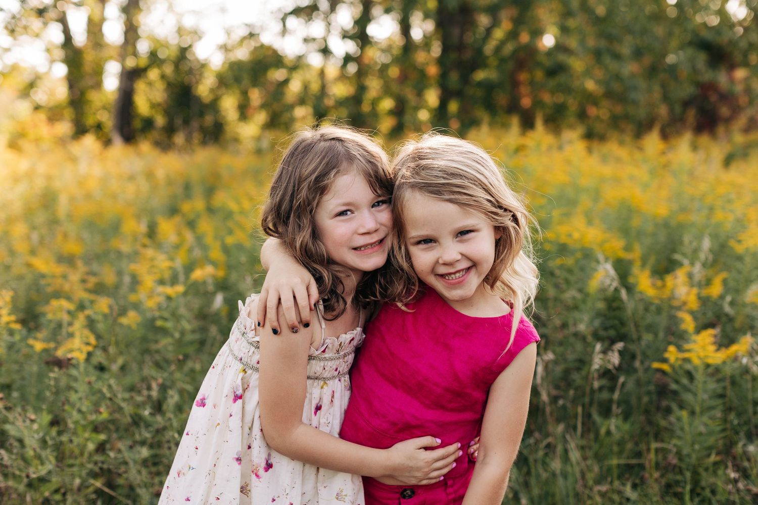 Two young girls hug each other warmly while standing in a field of yellow wildflowers at sunset.
