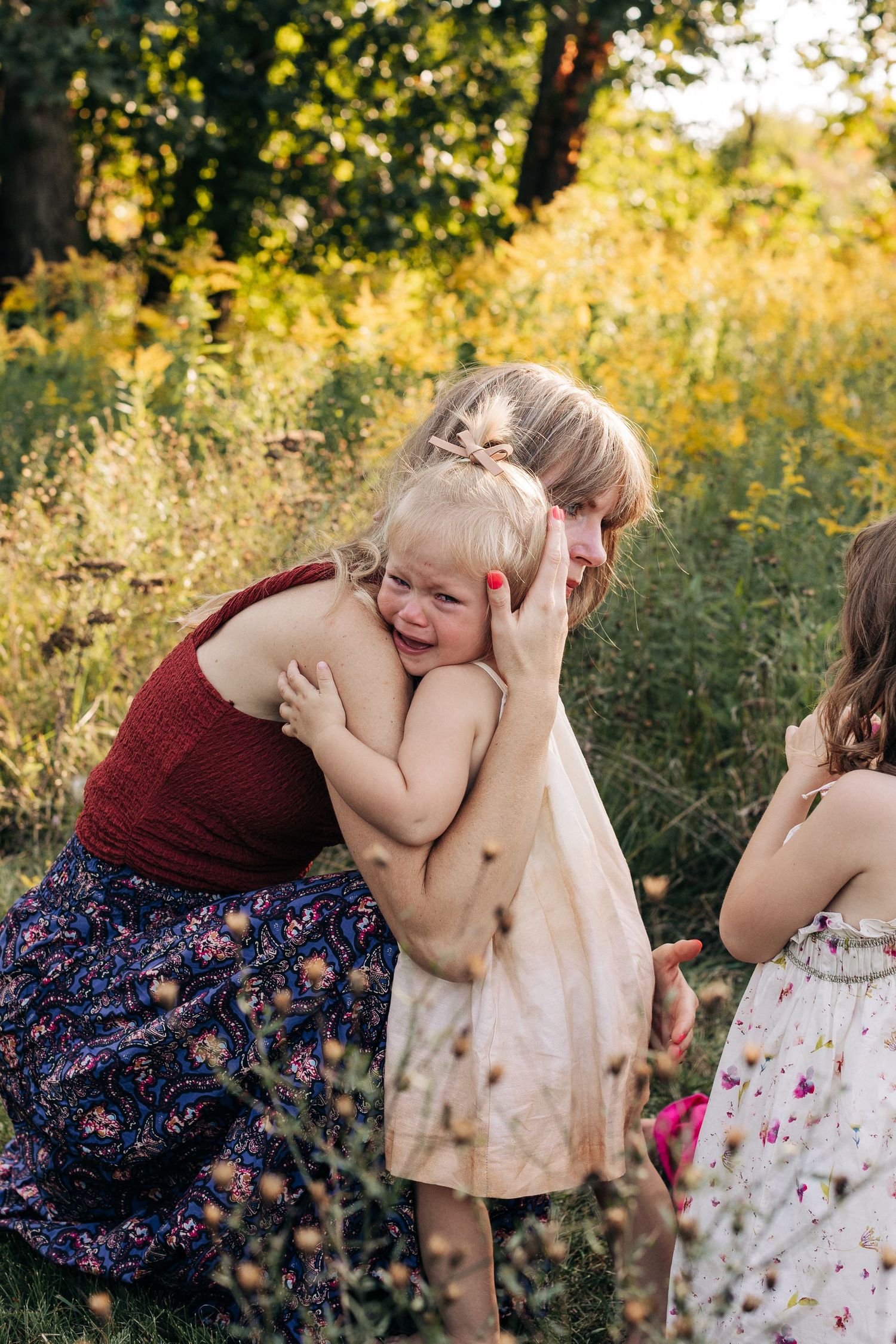 Mother in red top hugging crying toddler girl in beige dress outdoors in green meadow.