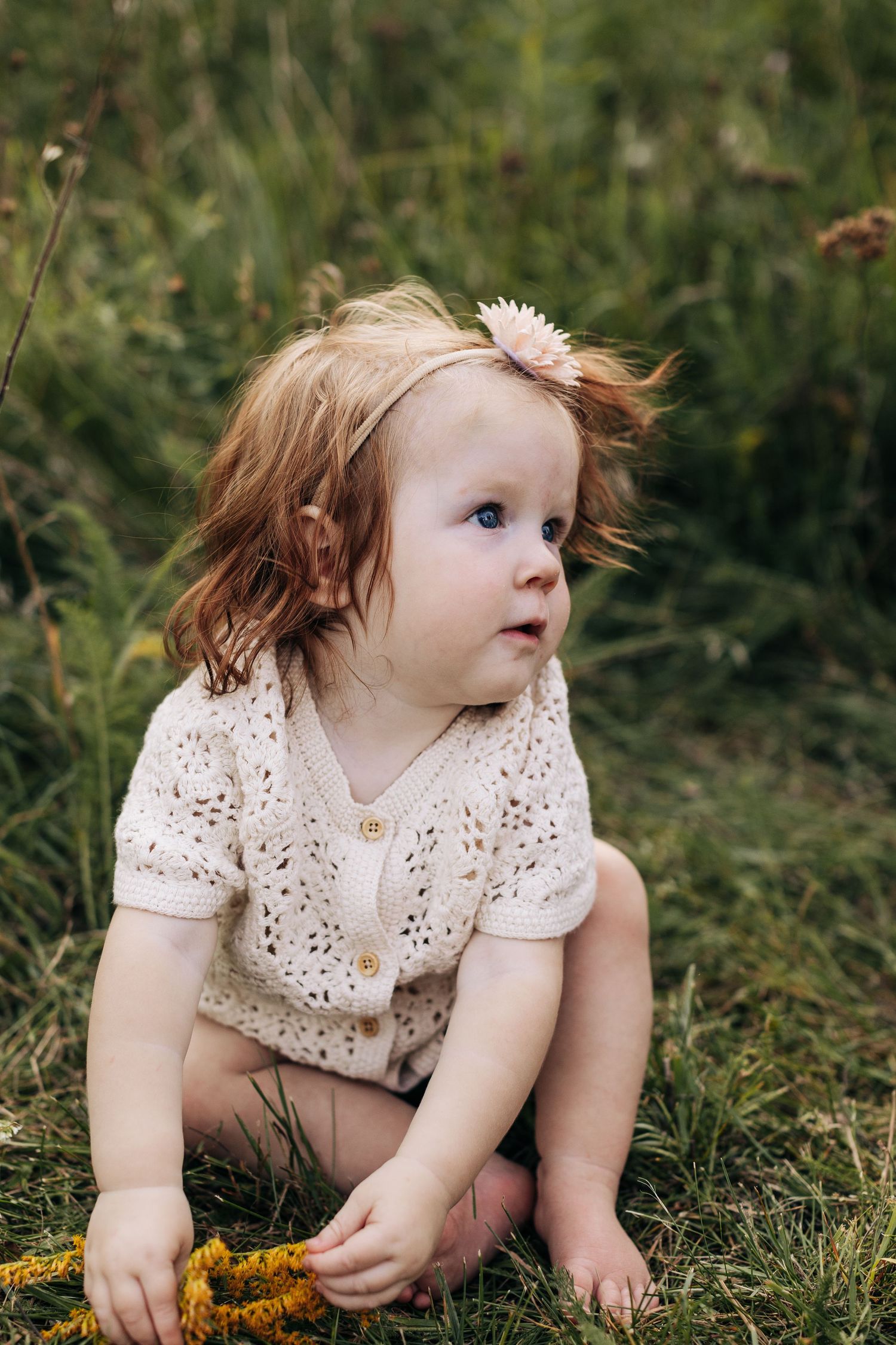 Adorable baby girl with pink flower headband sitting in grass holding yellow wildflowers.