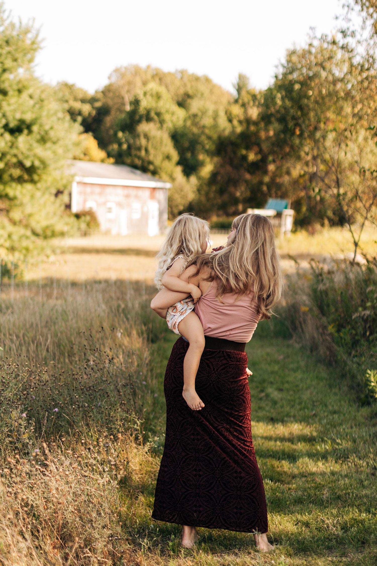 Mother carrying young child while walking through a scenic rural path at golden hour.