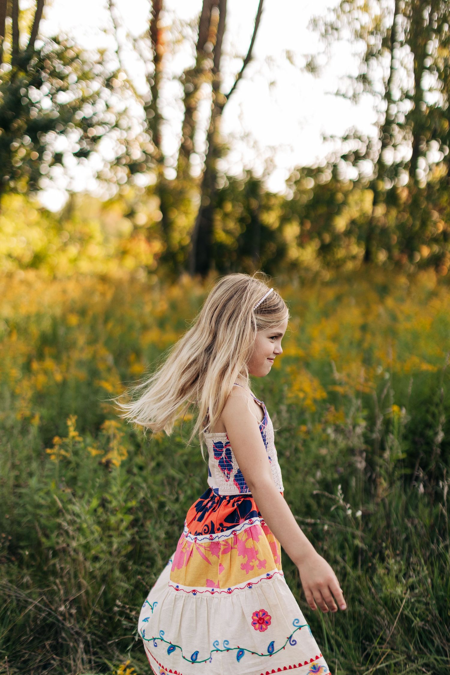 Young blonde girl in a colorful boho dress twirling in a golden wildflower meadow at sunset.