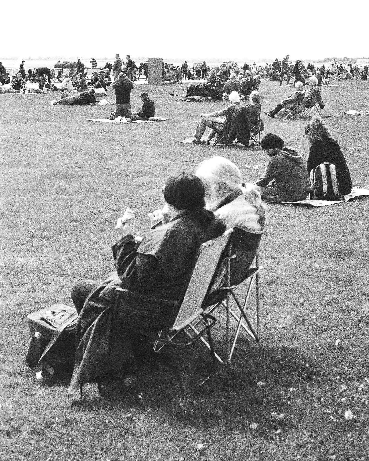 Black and white photo of people relaxing on grass at an outdoor event, with two seated in folding chairs in the foreground.
