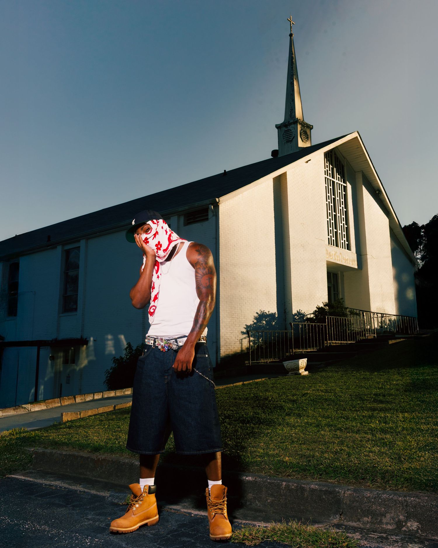 Young man in white tank top, denim shorts, and Timberland boots standing in front of a white church.
