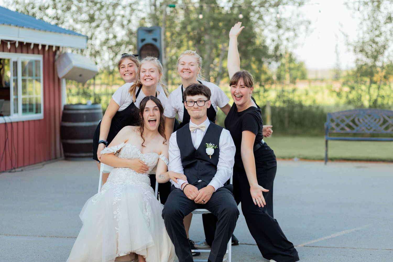 Wedding couple seated with four bridesmaids in black posing joyfully outdoors at a vineyard venue.