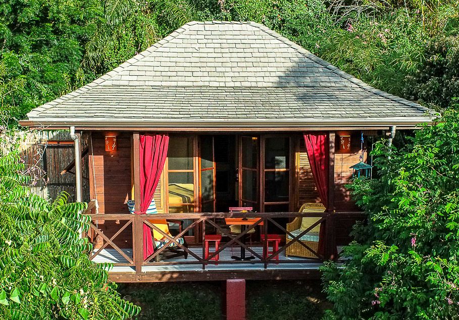 Cozy wooden bungalow with red curtains and a wraparound porch nestled among lush tropical greenery.