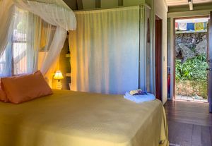 Cozy tropical bedroom with canopy bed, golden bedding, warm lamp, and open door revealing lush garden view.