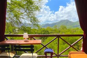 Balcony table with lush green mountain views under blue sky, framed by red curtains and wooden railing.