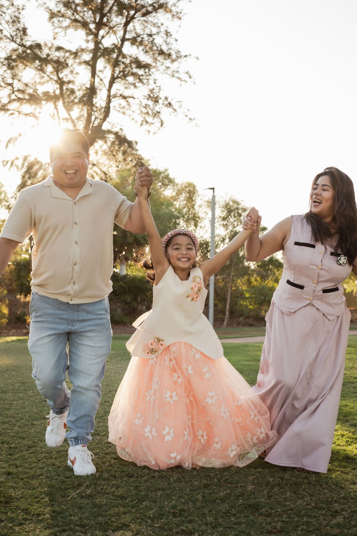 A joyful family of three holds hands and walks through a sunlit park, with a young girl in a pink floral dress.