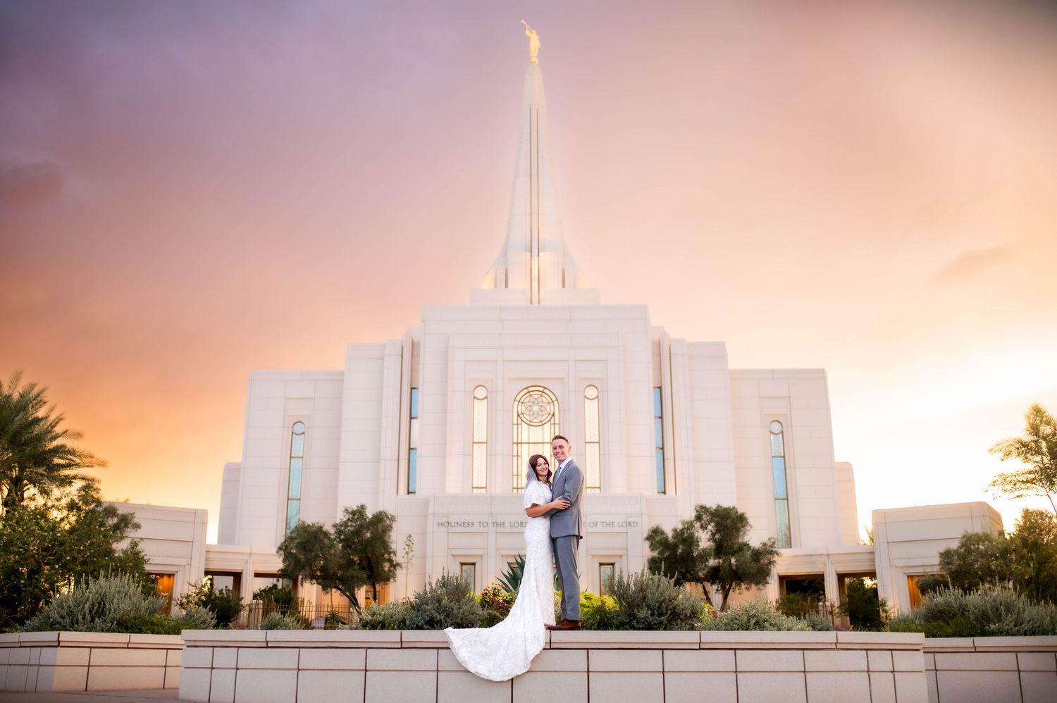 Bride and groom embracing before stunning white LDS temple under dramatic colorful sunset sky.